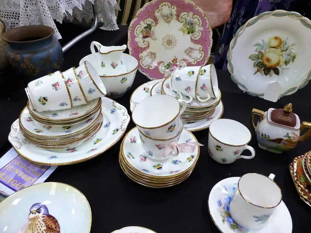 Een display met oude porseleinen theekopjes, schoteltjes en borden met bloemmotieven en gouden randjes, op een tafel naast antiek uit Ardingly, een decoratieve theepot, een vaas en andere keramische voorwerpen.