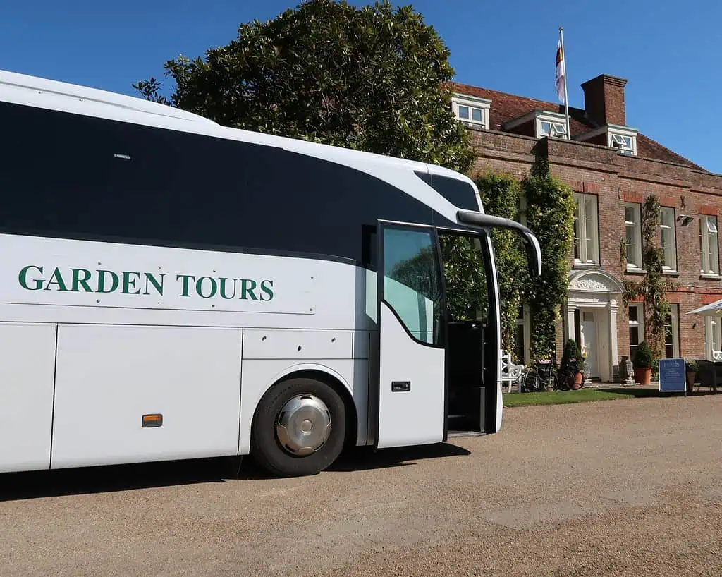 Een witte tourbus met het opschrift GARDEN TOURS staat geparkeerd voor een groot bakstenen landhuis, dat doet denken aan locaties die te zien zijn op de Chelsea Flower Show, met een vlag op het dak en weelderig groen onder een strakblauwe hemel.
