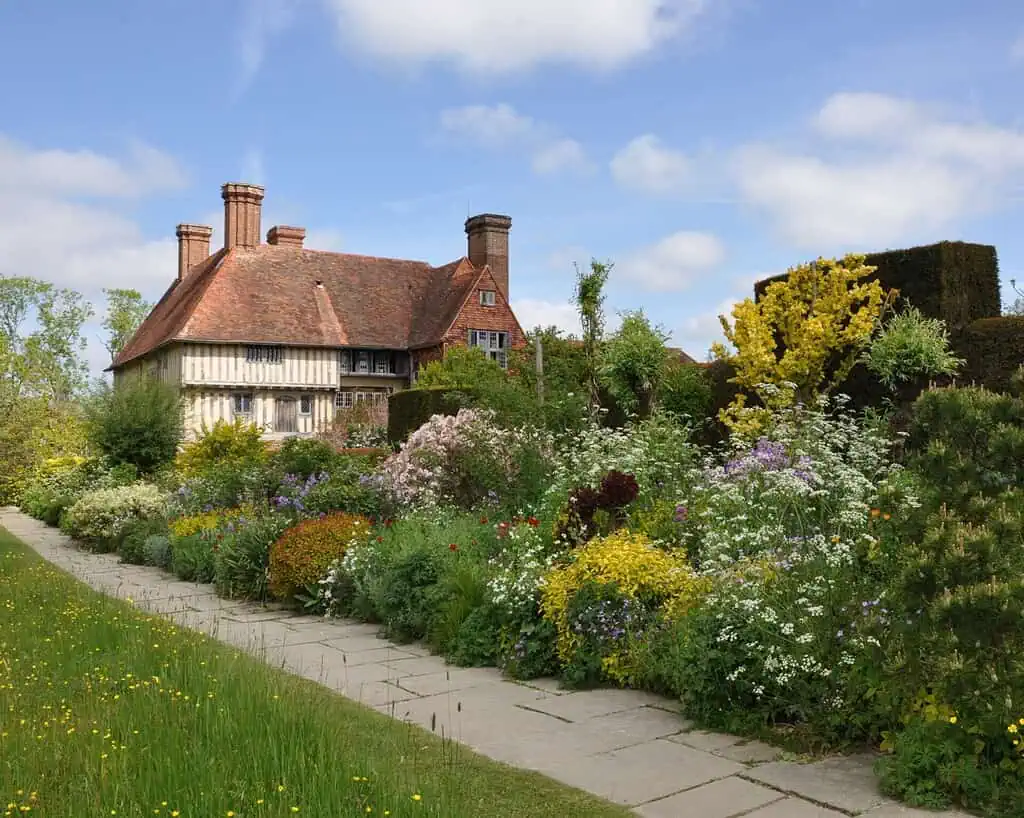 Een traditioneel Engels landhuis met een rood pannendak staat achter een weelderige, kleurrijke tuin geïnspireerd door de Chelsea Flower Show, vol bloeiende bloemen, groene struiken en een stenen pad onder een gedeeltelijk bewolkte blauwe hemel.