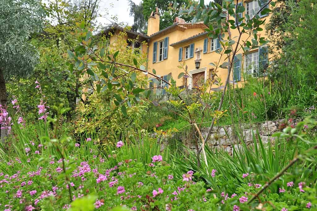 Een geel huis met groene luiken wordt omringd door weelderig groen en bloeiende roze bloemen in een levendige tuin. Het tafereel ziet er vredig en uitnodigend uit.