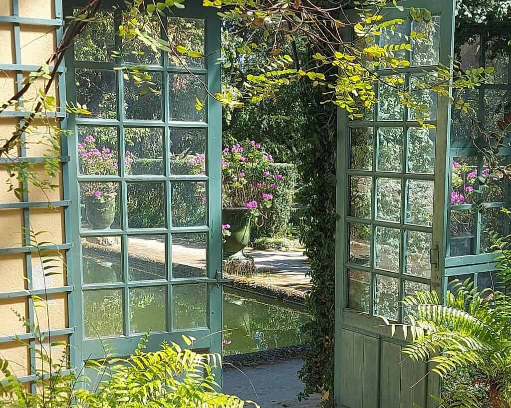 Zonlicht stroomt door open blauwe openslaande deuren en onthult een weelderige tuin met roze bloemen in plantenbakken, bladerrijke wijnranken en een rustige vijver, waardoor een sereen en uitnodigend buitenbeeld ontstaat.