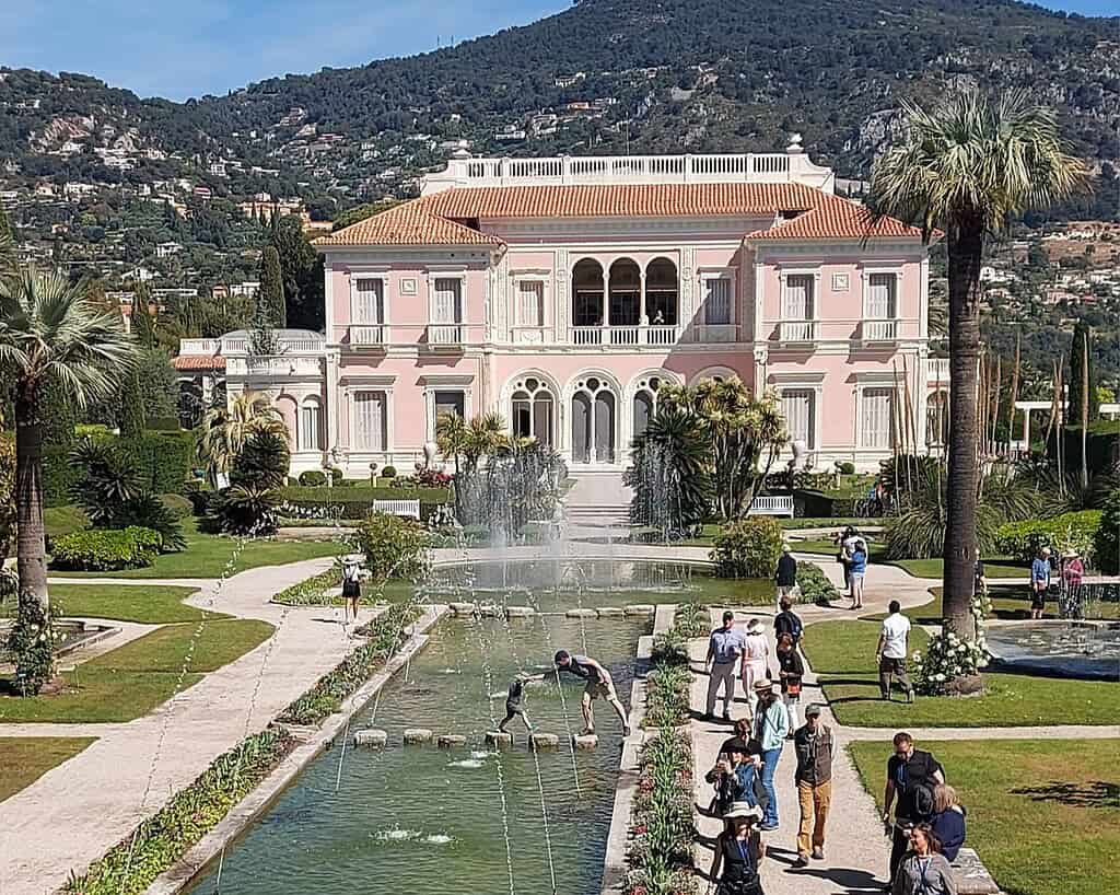 Een grote roze villa met gewelfde ramen ligt achter een formele tuin met fonteinen en palmbomen; bezoekers lopen en kruisen stenen trappen over een spiegelend zwembad, met groene heuvels op de achtergrond.