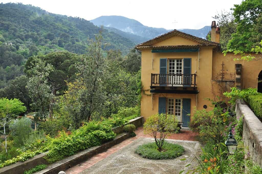 Een geel huis met blauwe luiken staat naast een weelderige, groene tuin en bomen, tegen beboste bergen onder een bewolkte hemel. Een stenen pad leidt naar de ingang.
