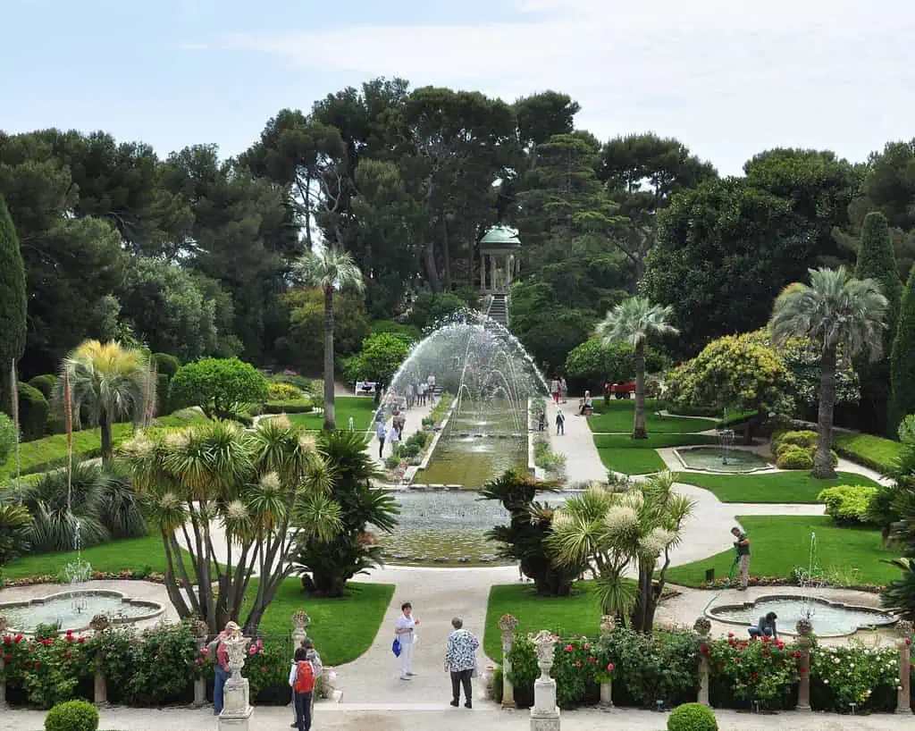 Een formele tuin met palmbomen, gemanicuurde gazons en symmetrische bloembedden, met een lange rechthoekige vijver met fonteinen die waterbogen spuiten en mensen die over paden lopen.