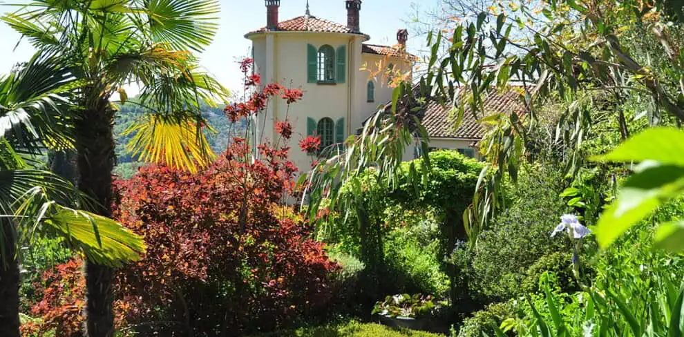 Een hoge villa met groene luiken en een rood pannendak staat achter weelderige, kleurrijke tuinen vol palmbomen, levendig rood en groen gebladerte en bloeiende bloemen op een zonnige dag.