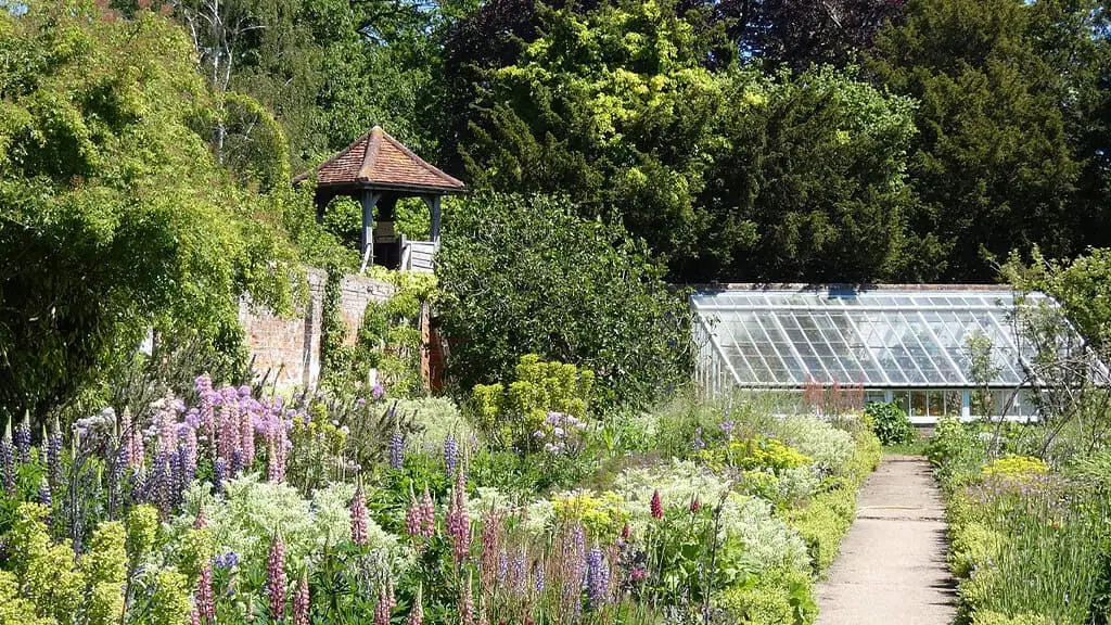 Een tuin met kleurrijke bloemen, een pad dat door weelderig groen kronkelt, een glazen kas en een klein houten prieel omringd door bomen vieren de schoonheid van de natuur en het plezier van groei onder helder zonlicht.