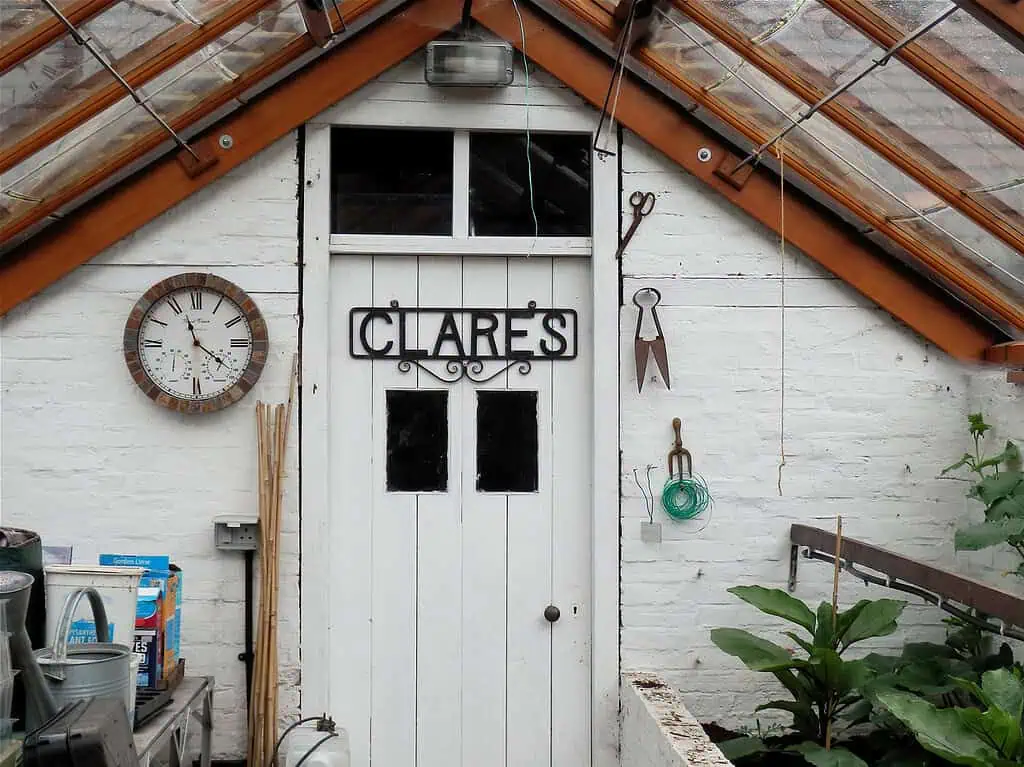 Witte deur met CLARES teken op de muur van een serre. Een klok, een tuinschaar en opgerold groen touw hangen aan de witte bakstenen muur. Tuinbenodigdheden en weelderige planten tonen de groei & bloei van de natuur onder het glazen dak.