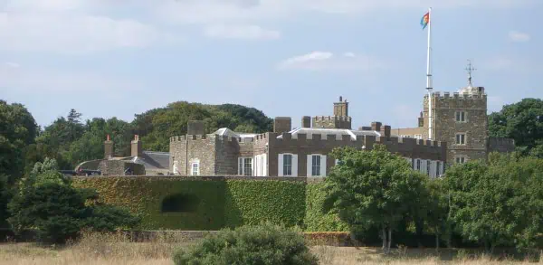 Een historisch stenen kasteel met wit omrande ramen en een hoge vlaggenmast wappert met een vlag, omringd door bomen en een met klimop begroeide tuin onder een gedeeltelijk bewolkte hemel.