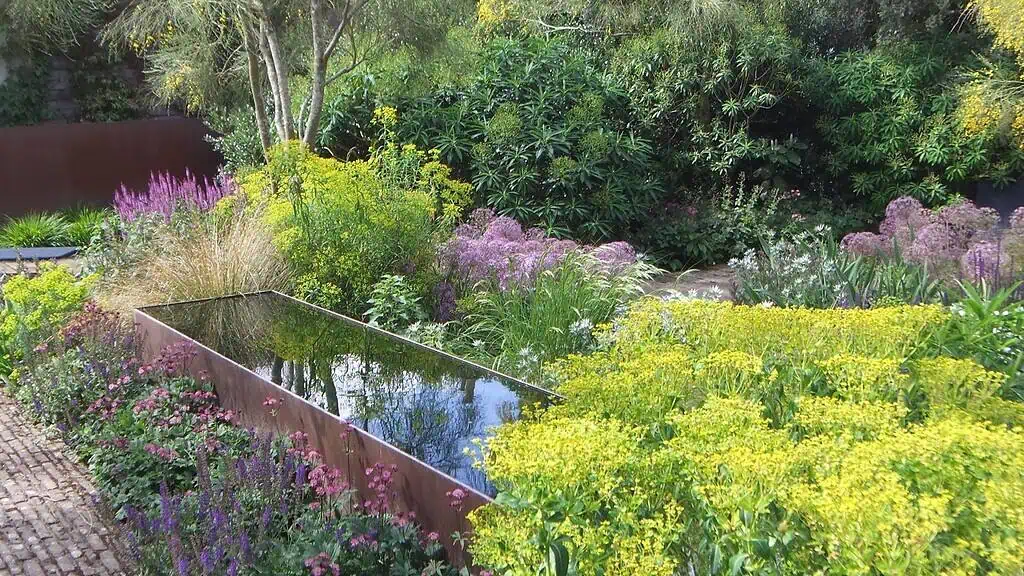Een rechthoekige spiegelende waterpartij ligt tussen weelderige, kleurrijke tuinplanten en wilde bloemen in een Groei & Bloei tuin, omringd door groene struiken en bomen onder natuurlijk daglicht.
