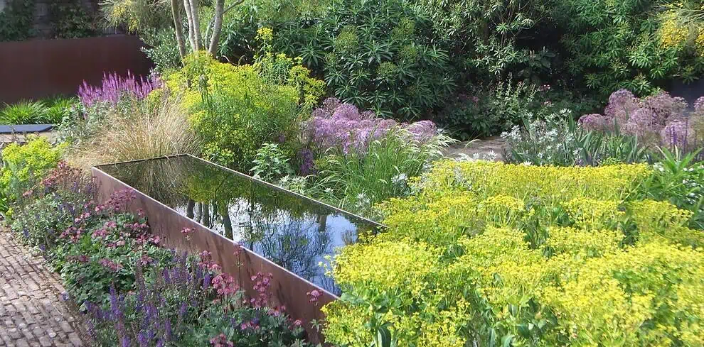 Een rechthoekige spiegelende waterpartij ligt tussen weelderige, kleurrijke tuinplanten en wilde bloemen in een Groei & Bloei tuin, omringd door groene struiken en bomen onder natuurlijk daglicht.