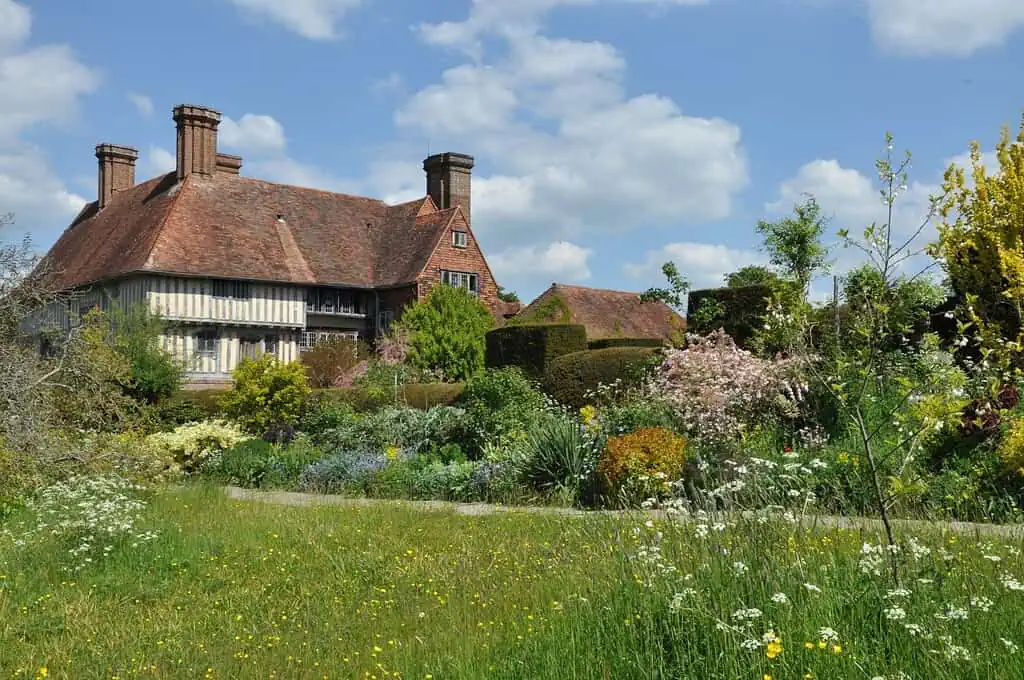 Een historisch huis met hoge schoorstenen en een steil pannendak staat achter een weelderige tuin vol wilde bloemen, struiken en bomen, waar de groei & bloei van de natuur te zien is onder een blauwe hemel met verspreide wolken.
