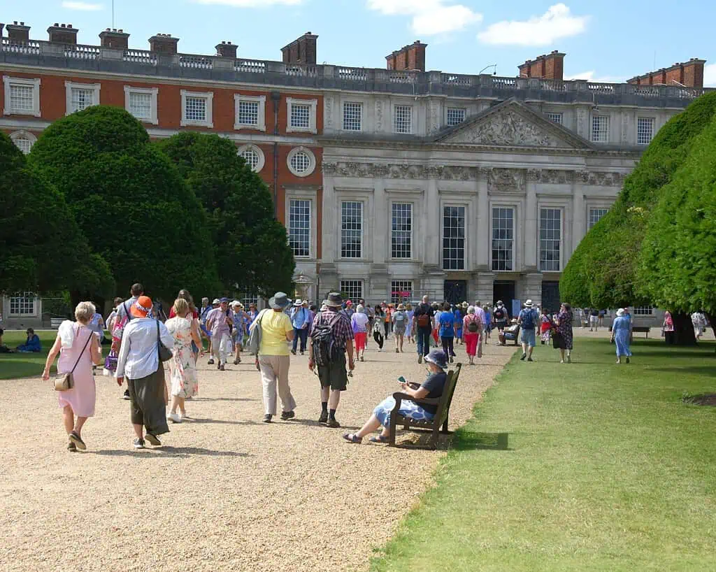 Bezoekers lopen over een grindpad naar een groots historisch gebouw met grote ramen en zuilen, omzoomd door de verzorgde koninklijke tuinen en gesnoeide bomen op een zonnige dag. Een persoon zit op een bankje op de voorgrond.