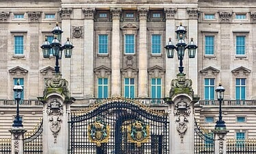 De afbeelding toont de sierlijke voorpoorten en gevel van Buckingham Palace, een van de beroemdste koninklijke paleizen, met zuilen, decoratief ijzerwerk en grote ramen onder een blauwe hemel.