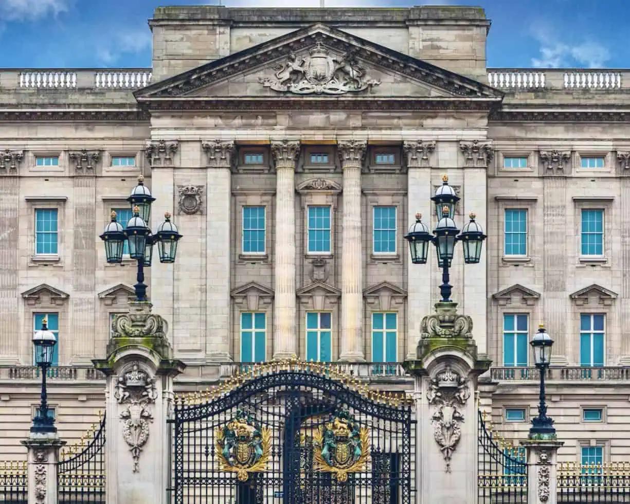 De afbeelding toont de sierlijke voorpoorten en gevel van Buckingham Palace, een van de beroemdste koninklijke paleizen, met zuilen, decoratief ijzerwerk en grote ramen onder een blauwe hemel.