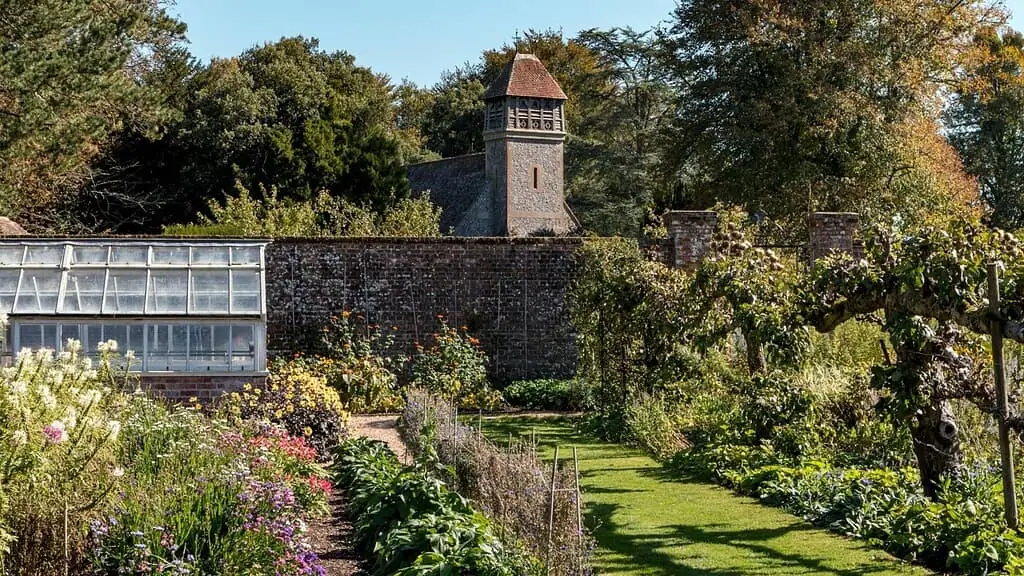 Een weelderige tuin met kleurrijke bloemen en planten omzoomd door een bakstenen muur, een glazen kas aan de linkerkant en een stenen gebouw met toren en pannendak op de achtergrond, omringd door bomen - perfect voor een Groei & Bloei moestuinreis.