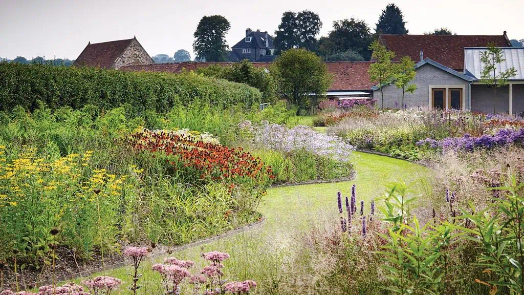 Een weelderige, kleurrijke tuin met een kronkelend graspad loopt door levendige bloemperken en een bloeiende moestuin. Boerderijen en huizen met rode daken op de achtergrond worden omzoomd door hagen en bomen onder een bewolkte hemel, perfect voor een Groei & Bloei moestuinreis.