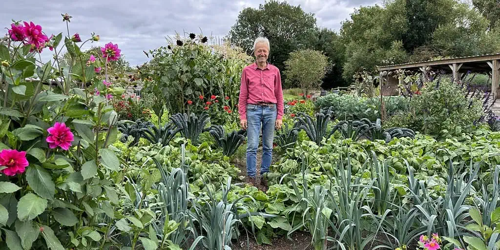 Een man in een rood shirt en spijkerbroek staat midden in een weelderige Groei & Bloei moestuin, omringd door bladgroen en bloemen, met bomen en een bewolkte lucht op de achtergrond - de essentie van zijn Moestuinreis.