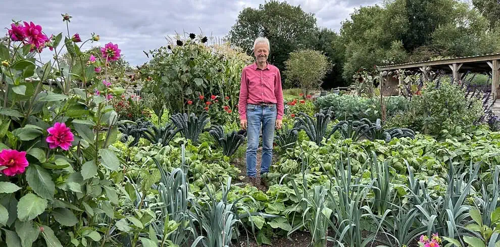 Een man in een rood shirt en spijkerbroek staat midden in een weelderige Groei & Bloei moestuin, omringd door bladgroen en bloemen, met bomen en een bewolkte lucht op de achtergrond - de essentie van zijn Moestuinreis.