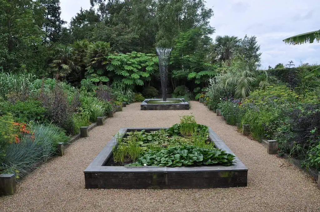 Een grindpad loopt tussen twee rechthoekige vijvers omringd door weelderige tuinplanten; de verste vijver heeft een hoge waterfontein die omhoog spuit en bomen en groen vullen de achtergrond.