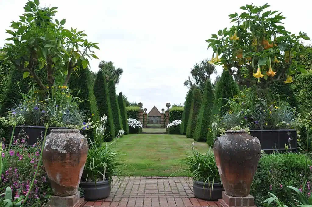 Een formele tuin met grote terracotta potten en bloeiende planten die een stenen pad flankeren, dat leidt naar een keurig gemaaid gazon omzoomd door gesnoeide heggen en eindigt bij een klein paviljoen in de verte.