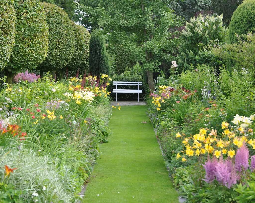Een wit bankje staat aan het einde van een weelderig groen pad met levendige bloemen en netjes gesnoeide struiken in een vredige tuin.